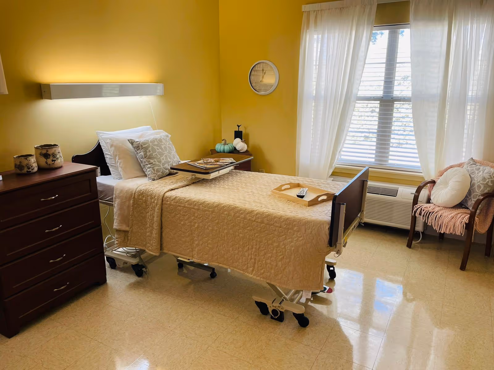 A bright and cozy room with a single hospital-style bed covered in beige bedding. The bed has a tray table with magazines and a wooden tray with a remote control. There is a wooden dresser with decorative pots on the left side, a small side table with decorative pumpkins, and a wooden chair with cushions and a pink throw blanket near a window with white curtains. The walls are painted yellow, and the floor is tiled.