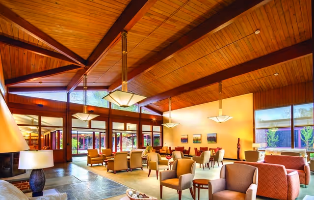 Spacious senior living lounge with a high wood-beamed ceiling, pendant lights, multiple seating groupings, and large windows.