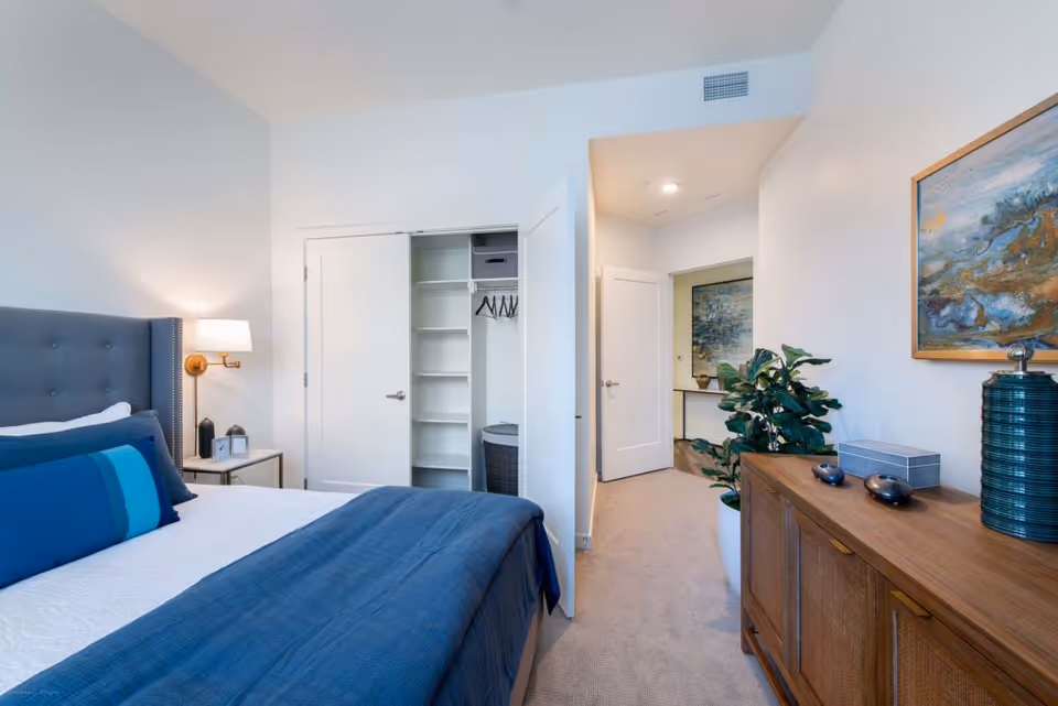 A modern bedroom with a neatly made bed featuring blue and white bedding, a gray upholstered headboard, and a bedside table with a lamp and decorative items. The room has an open closet with shelves and hangers, a wooden dresser with decorative objects and a large plant beside it. A hallway with artwork on the wall is visible in the background.