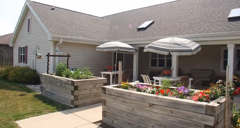 Outdoor patio area at Lakepoint Villa Assisted Living featuring raised wooden garden beds with colorful flowers and plants, two striped umbrellas shading tables and chairs, and a building with beige siding and a sloped roof with skylights in the background.