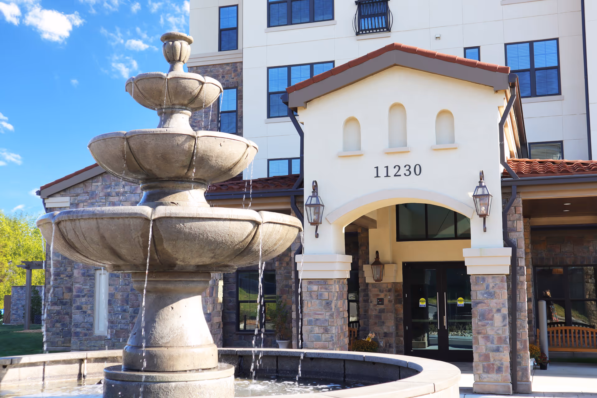 Stone water fountain with multiple tiers in front of the entrance to a senior living facility building with stone and stucco exterior, arched doorway, and the number 11230 above the entrance.