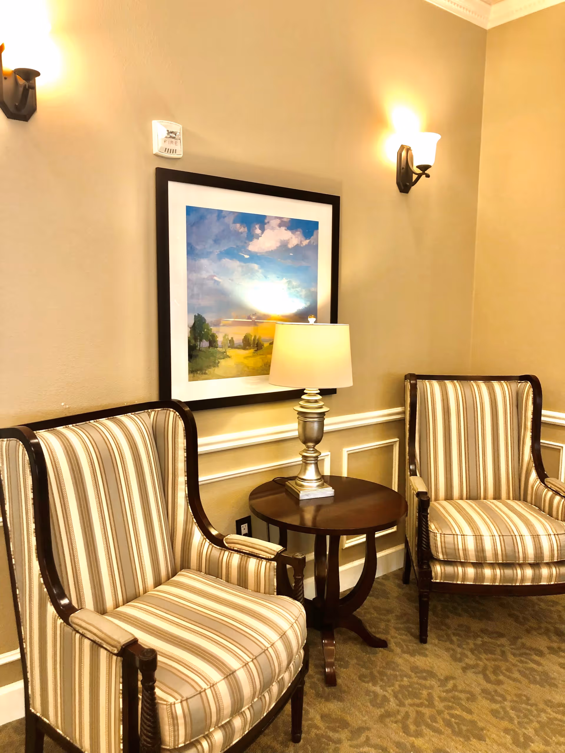 A cozy seating area with two striped upholstered armchairs separated by a round wooden side table with a silver lamp on it. Above the table hangs a framed landscape painting featuring a blue sky with clouds and a green field. The walls are beige with white trim, and two wall sconces provide warm lighting.