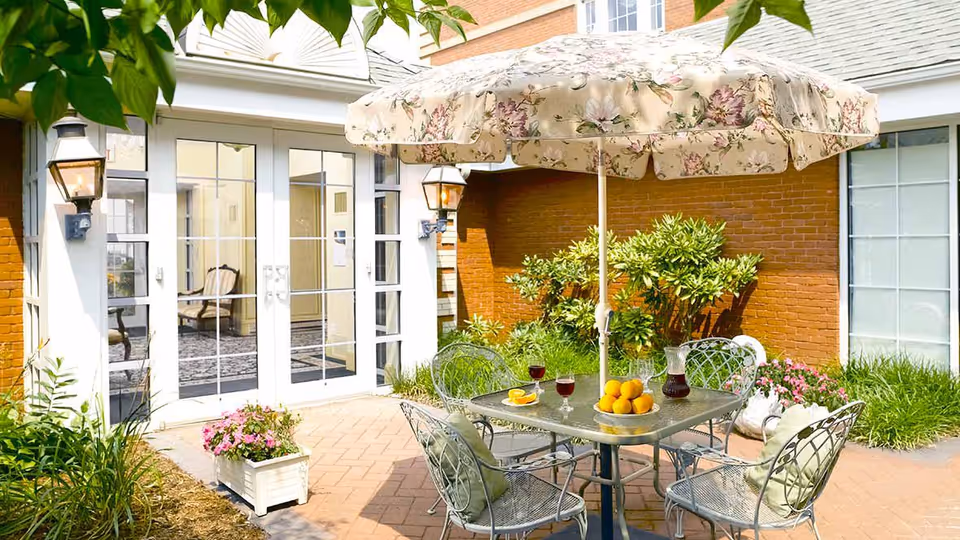 Outdoor patio area with a square metal table and four metal chairs with cushions. The table has a floral-patterned umbrella providing shade, a pitcher of red drink, four glasses filled with the drink, a plate with orange slices, and a bowl of oranges. The patio is surrounded by plants and flowers, with a brick wall and glass doors leading inside the building.