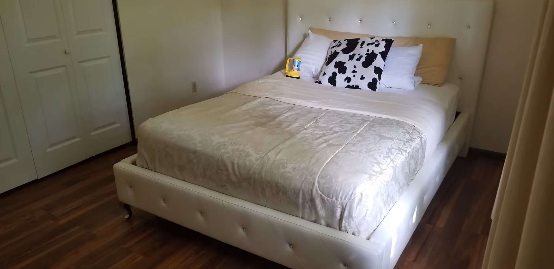 A neatly made bed with a tufted white headboard, patterned comforter and cow-print pillow in a simple bedroom with hardwood floors and closet doors.