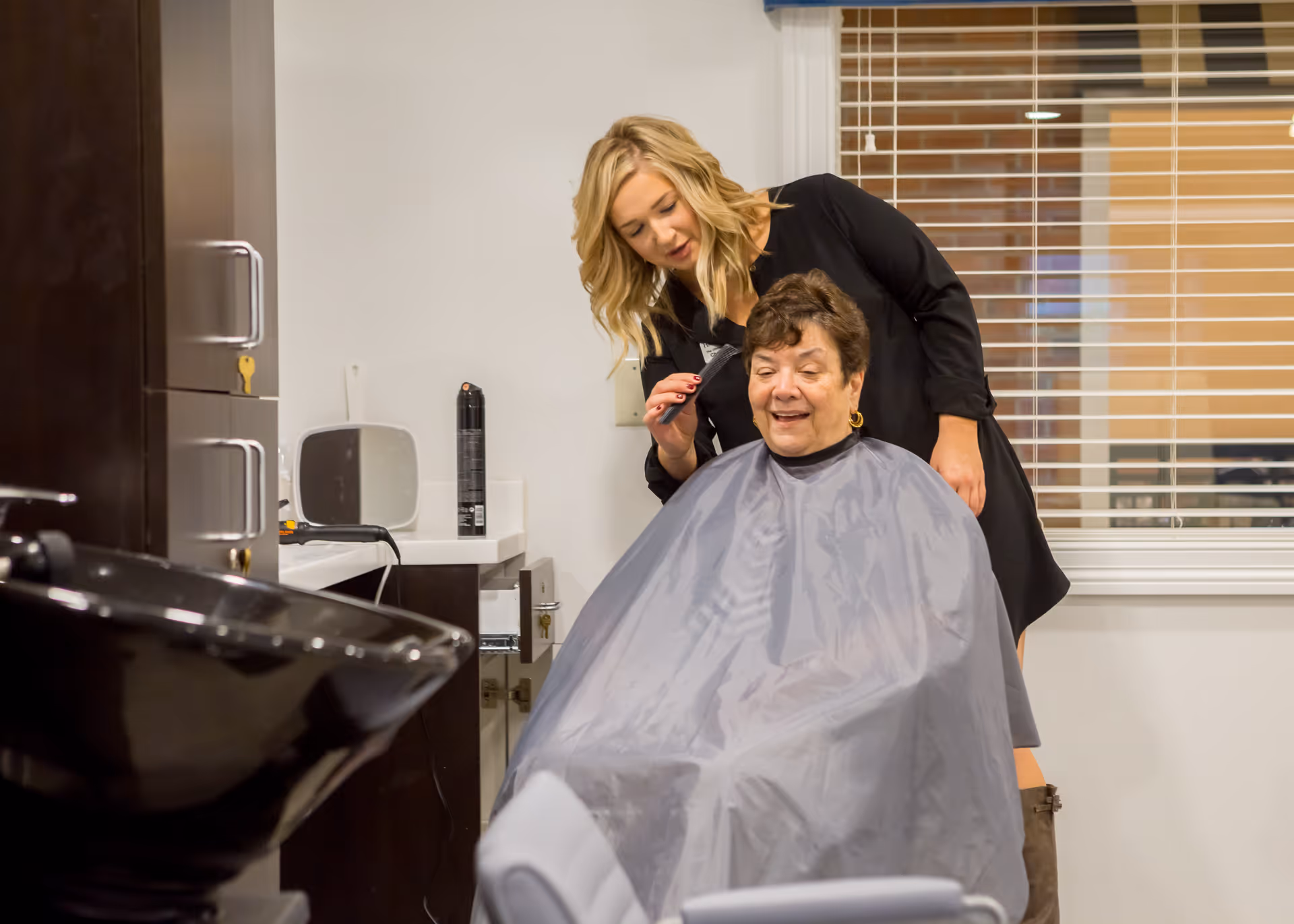 A hairstylist is cutting the hair of an elderly woman who is seated and covered with a gray cape in a salon area with a black sink and cabinets in the background.