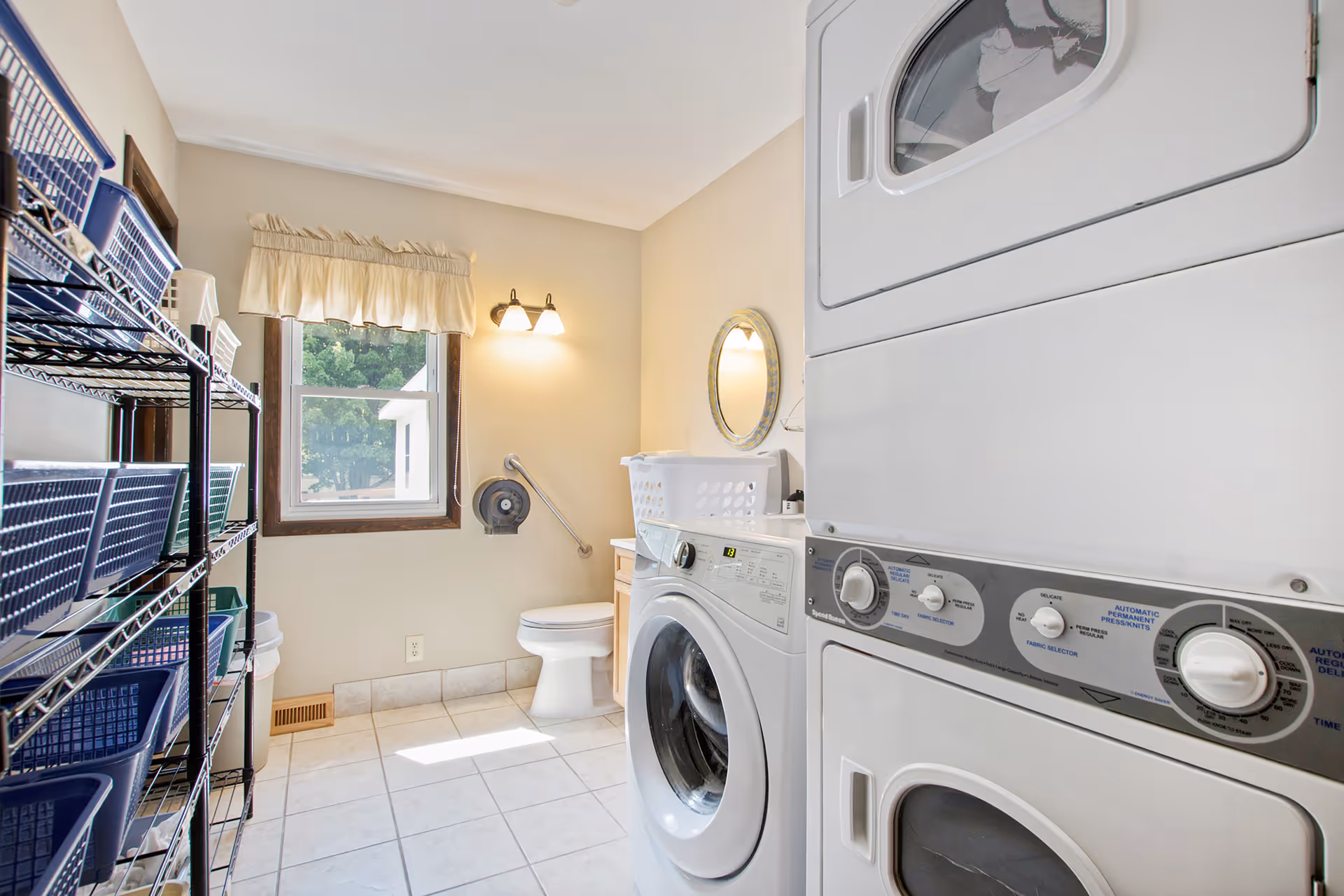 A bright laundry room with a stacked washer and dryer on the right side, a front-loading washing machine next to them, and a white laundry basket on top. On the left side, there is a metal shelving unit with multiple blue baskets. A window with a beige valance lets in natural light, and a toilet is visible in the corner under a wall-mounted light fixture and a round mirror.