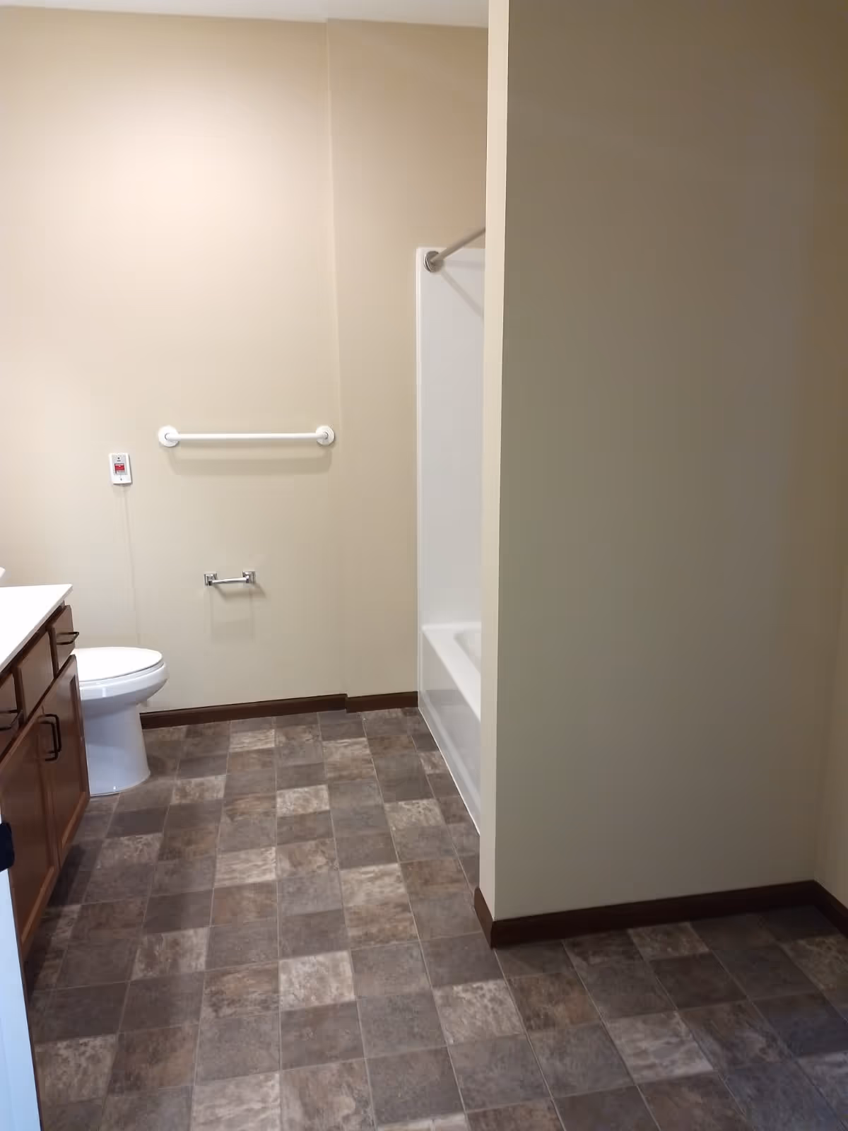 A clean bathroom with beige walls and a tiled floor. The room features a white toilet, a wooden cabinet with a countertop, a bathtub with a shower curtain rod, a white grab bar on the wall, and a toilet paper holder without a roll.