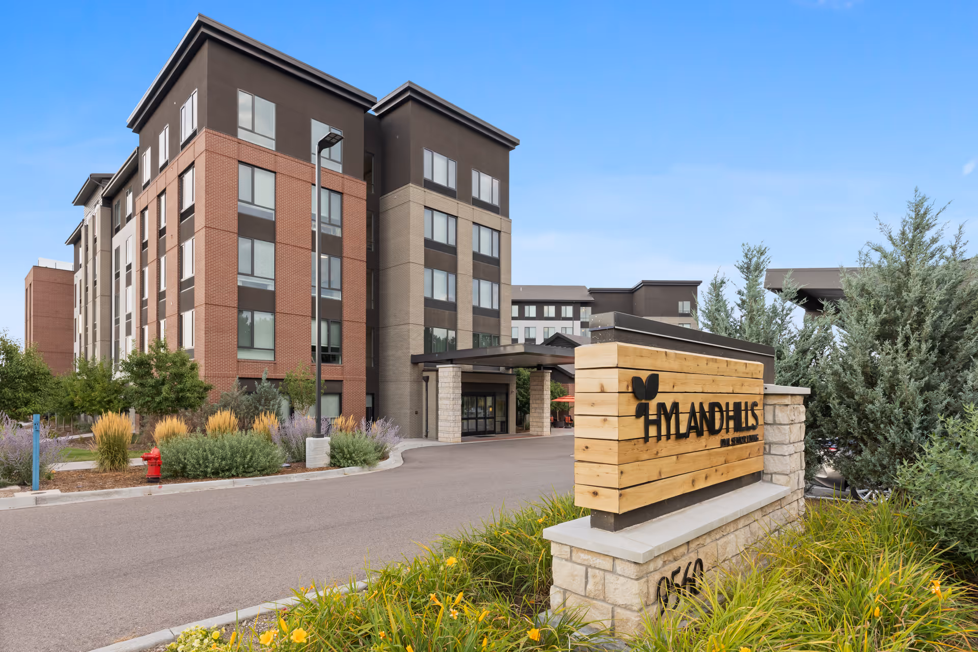 Exterior view of Hyland Hills Senior Living facility showing a multi-story building with large windows, landscaped greenery, and a wooden sign with the facility's name near the entrance driveway.