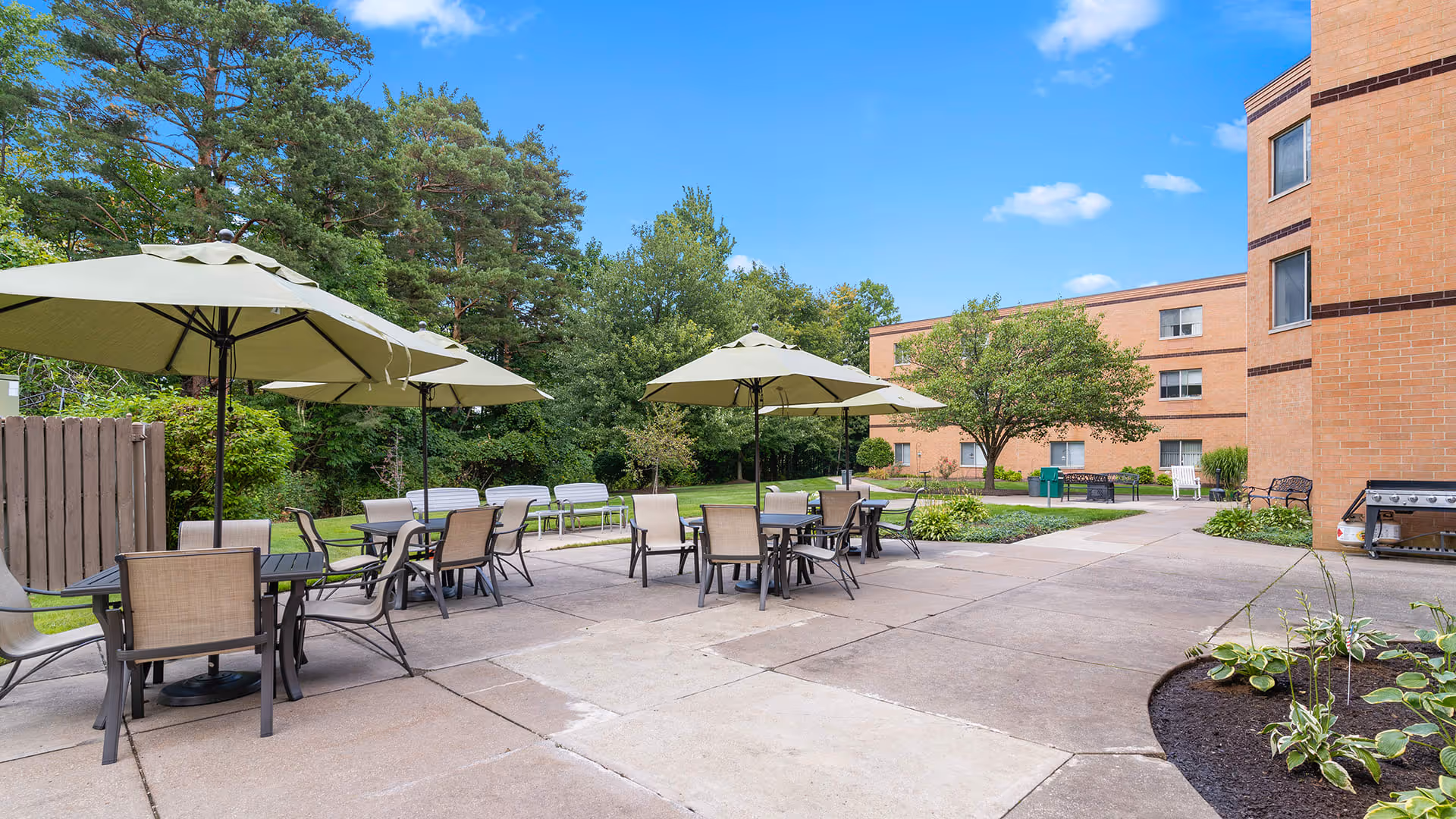 Outdoor patio area at Holiday Niagara Village with several tables and chairs under large beige umbrellas, surrounded by greenery and trees, with a brick building in the background under a clear blue sky.