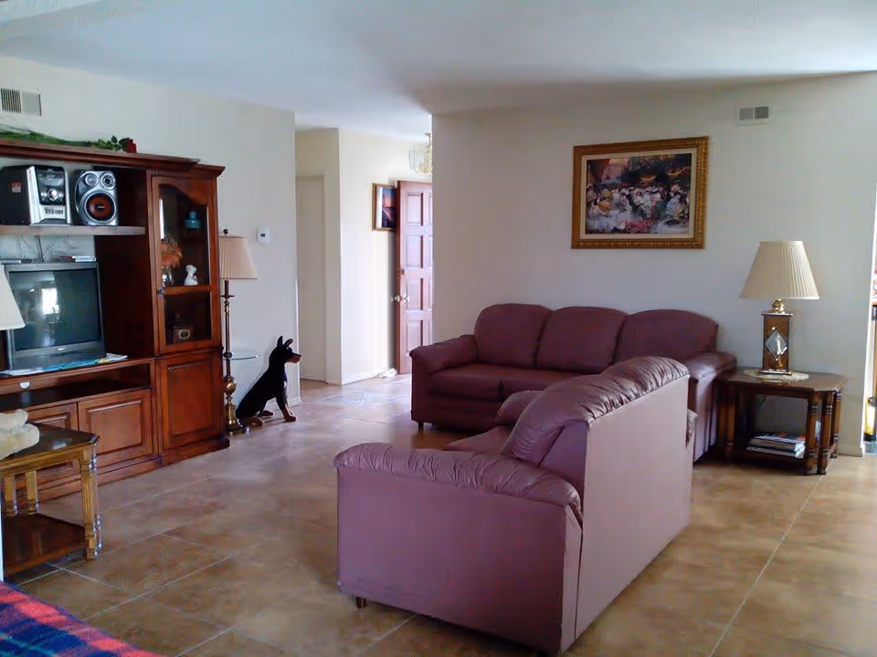 A living room with two mauve leather sofas arranged around a wooden side table with a lamp. There is a wooden entertainment center with a TV, stereo system, and decorative items. A framed painting hangs on the wall above one sofa. The floor is tiled, and there is a doorway leading to another room.