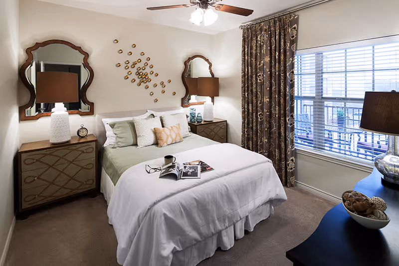 A neatly made bed with white and green bedding in a bedroom. There are two matching bedside tables with lamps and decorative mirrors above them. A decorative wall art piece is above the bed. A window with patterned curtains lets in natural light, and a dark dresser with a lamp and decorative bowl is on the right side of the room.