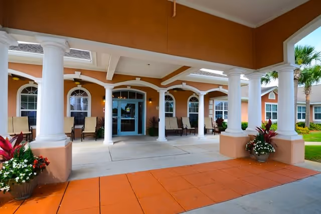 Entrance area of a senior living facility with white columns supporting a covered walkway. There are several beige chairs lined up against the building's peach-colored exterior walls with arched windows. Potted plants with flowers are placed near the columns and along the walkway. The entrance door is glass with a blue frame.