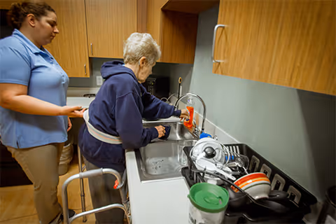 An elderly woman using a walker is washing her hands at a kitchen sink with assistance from a caregiver standing beside her in a room with wooden cabinets and a dish rack filled with dishes.
