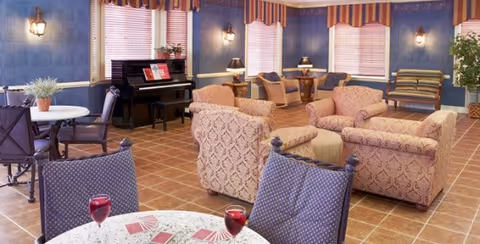 A spacious and well-lit living room area with patterned upholstered armchairs and sofas arranged around a small round table. There is a piano against the wall with sheet music on it, several small tables with lamps, and a striped bench near the windows. The room has tiled floors, blue walls, and windows with blinds and striped valances. A round table with chairs and two glasses of red drink is visible in the foreground.