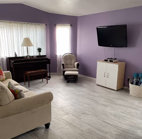 A cozy living room with light gray tiled flooring and purple walls. The room features a beige sofa with colorful pillows, a wooden piano with a lamp and small plant on top, a cushioned armchair with a matching footrest, a wall-mounted flat screen TV above a small cabinet, and a basket holding rolled-up mats or posters. Two windows with sheer white curtains allow natural light into the space.