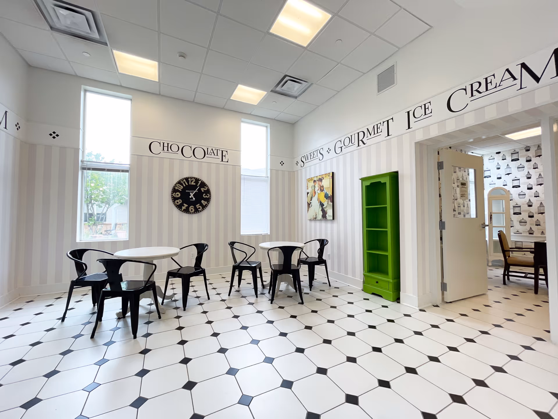 A bright room with white walls featuring vertical stripes and large windows letting in natural light. The walls have decorative text reading 'CHOCOLATE', 'SWEETS', 'GOURMET ICE CREAM'. The floor is tiled with a white and black geometric pattern. There are two small round white tables each surrounded by three black metal chairs. A black clock with white numbers hangs on the wall between the windows. A bright green shelving unit stands against the wall near an open doorway leading to another room with patterned wallpaper and additional seating.