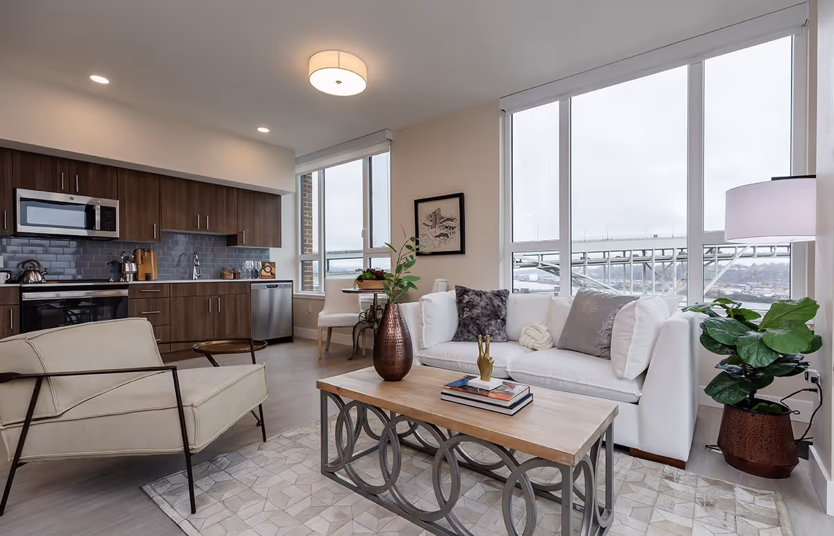 Modern open-concept living room and kitchen area with large windows showing a bridge outside. The living room features a white sofa with decorative pillows, a wooden coffee table with books and a vase, a beige armchair, a floor lamp, and a potted plant. The kitchen has dark wood cabinets, a microwave, oven, dishwasher, and a small dining table with chairs near the window.