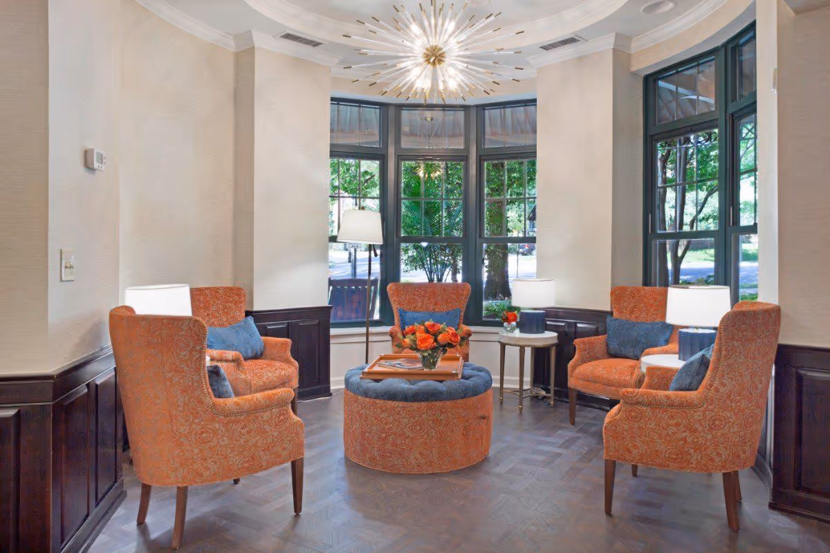 A cozy seating area with four orange upholstered armchairs arranged around a round ottoman and coffee table in front of large bay windows.