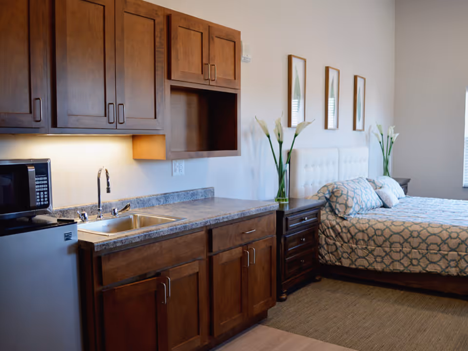 A studio apartment interior featuring a kitchenette with wooden cabinets, a small sink, a microwave, and a mini refrigerator. Adjacent to the kitchenette is a bed with a white tufted headboard, patterned bedding, two bedside tables with vases of white flowers, and three framed botanical prints on the wall.