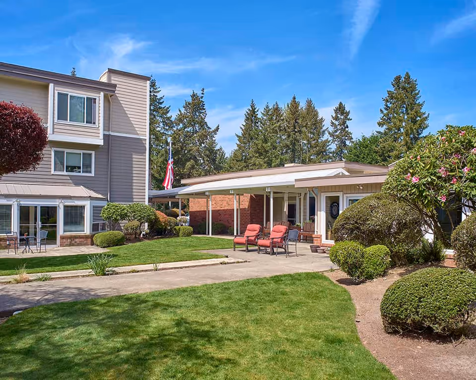 Outdoor view of Sagebrook Senior Living at Bellevue featuring a well-maintained lawn, trimmed bushes, and two buildings with seating areas under a clear blue sky.