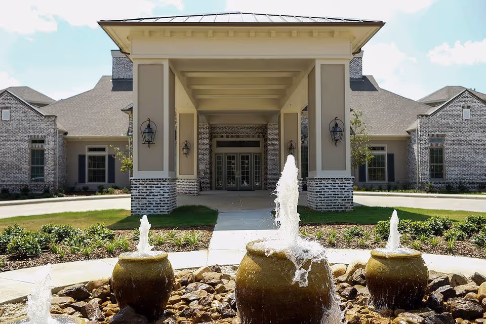 Front entrance of The Claiborne at McComb facility featuring a covered driveway with columns, brick exterior walls, and a decorative water fountain with three spouts in the foreground surrounded by landscaping.