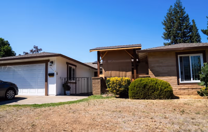 A single-story suburban home exterior with a garage, front yard, and shrubs.
