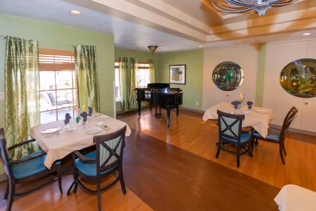 A dining room with wooden floors and light green walls featuring two tables set with white tablecloths, plates, glasses, and silverware. Each table has four chairs with blue cushions. There are large windows with green curtains letting in natural light. A black grand piano is positioned near the corner of the room, and two round mirrors are mounted on the far wall.