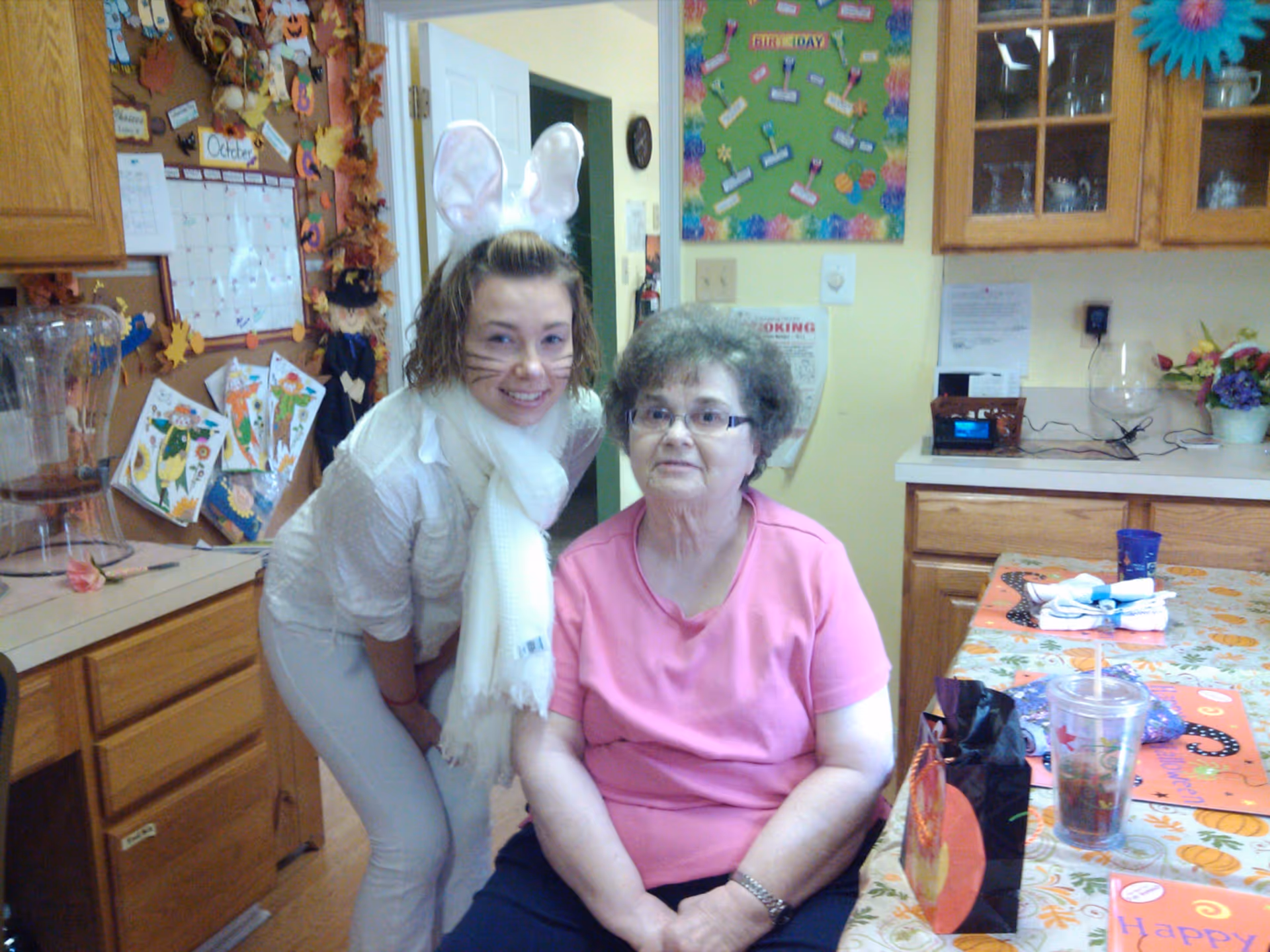 An elderly woman wearing a pink shirt sitting at a table with fall-themed decorations and a younger woman dressed in a white outfit with bunny ears and face paint standing beside her in a kitchen area.