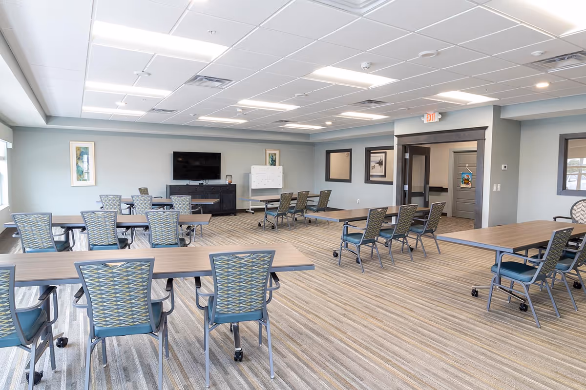 Spacious assisted-living common room with several tables and patterned chairs, a wall-mounted TV and a whiteboard.
