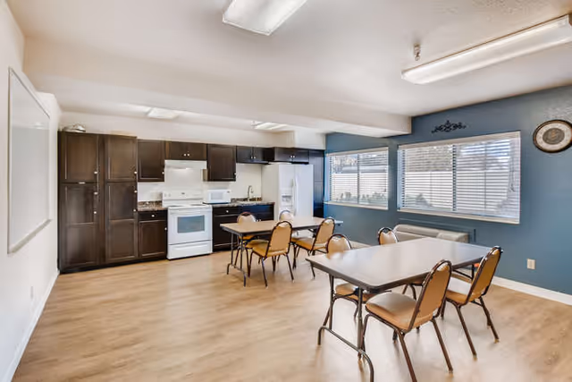 Communal dining room with tables and chairs and a kitchenette featuring dark cabinets, white appliances, and large windows.