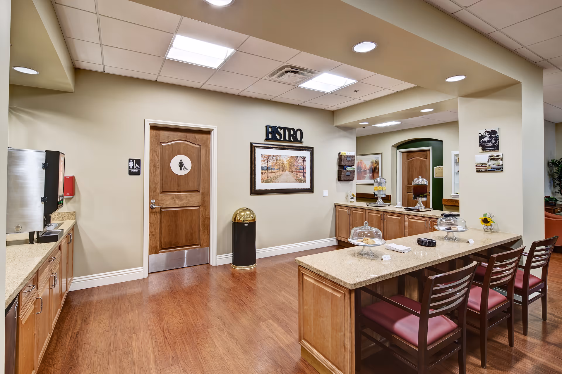 Interior view of a senior living facility's bistro area with wooden flooring, a countertop island with chairs, cabinets, beverage dispensers, and a restroom door marked with a women's restroom sign. The walls are decorated with framed pictures and a sign that reads 'BISTRO'.