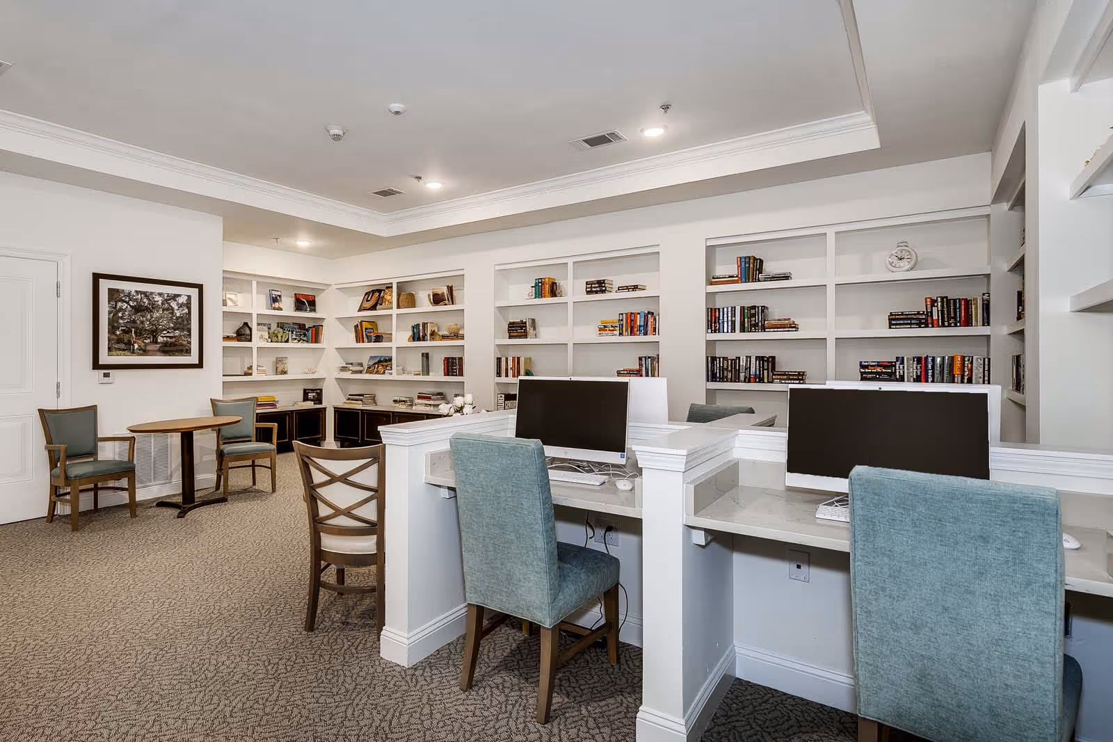A quiet library or computer room in a senior living facility with built-in white bookshelves filled with books and decorative items. There are two computer stations with monitors and teal upholstered chairs. In the background, there is a small round table with two wooden chairs and a framed picture on the wall.