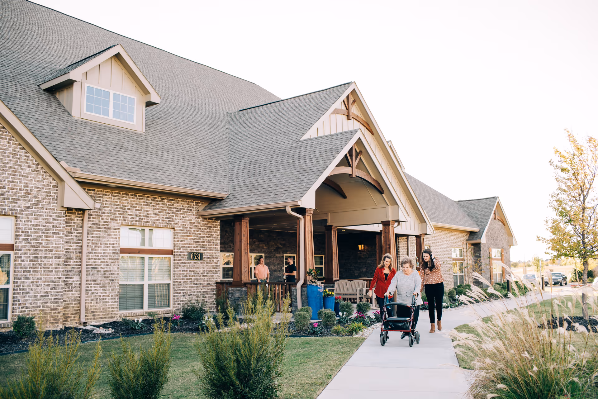 Exterior view of a brick assisted living facility with a large covered entrance. Two women are walking on the sidewalk, one using a walker, and two men are standing under the covered entrance talking. The building has multiple windows and a well-maintained garden with shrubs and flowers.
