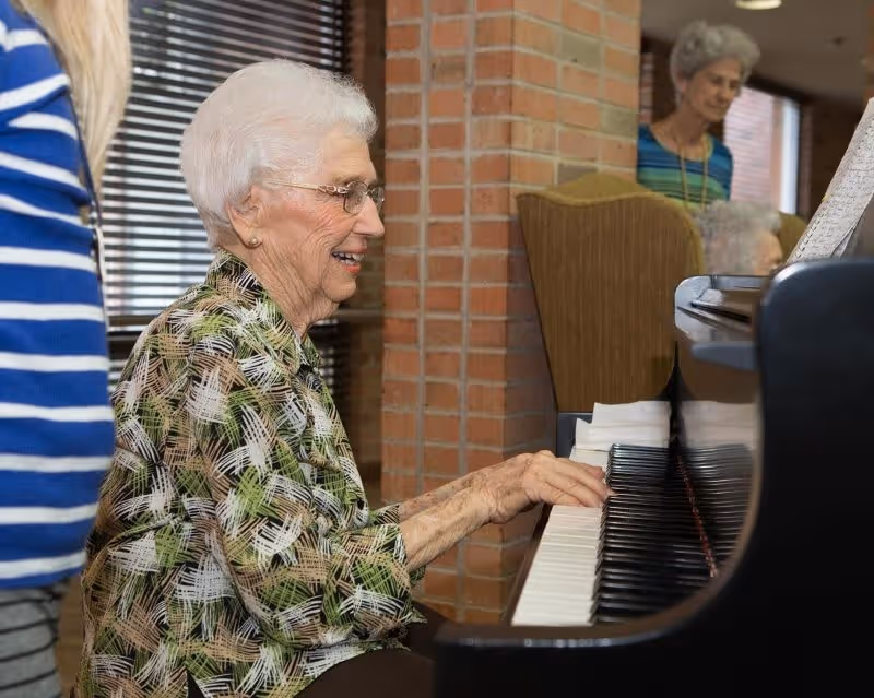 An elderly woman with white hair and glasses is playing a piano indoors. She is smiling and wearing a patterned blouse. In the background, another elderly woman is standing near a brick wall and window blinds, and a third person is partially visible sitting near the piano.
