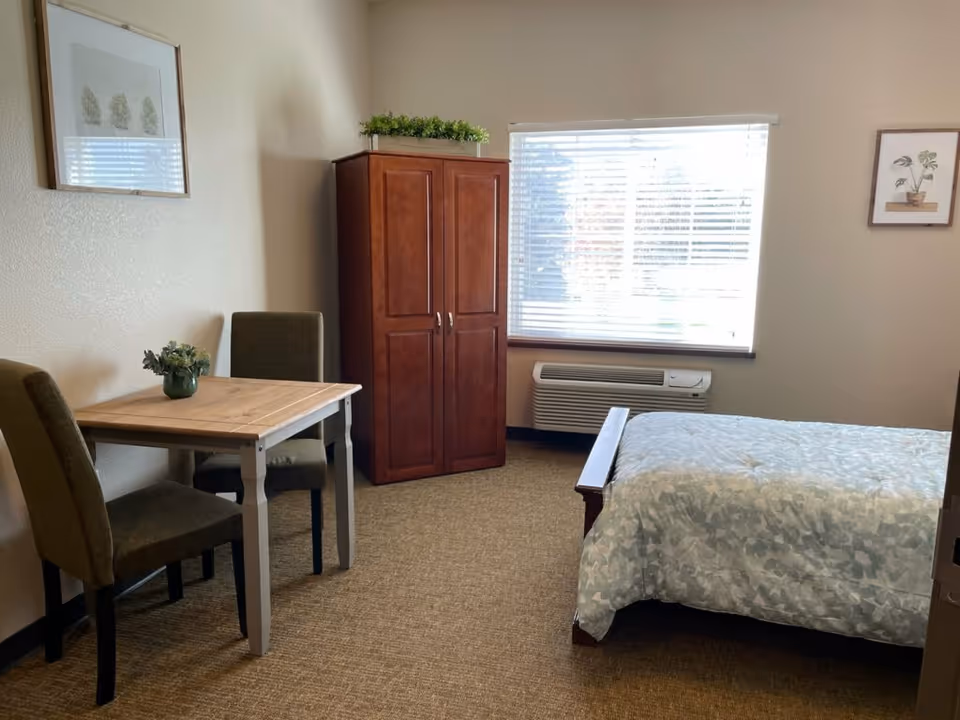 A cozy bedroom in an assisted living facility featuring a bed with a patterned comforter, a wooden wardrobe, a small table with two chairs, a window with blinds, and framed botanical artwork on the walls.