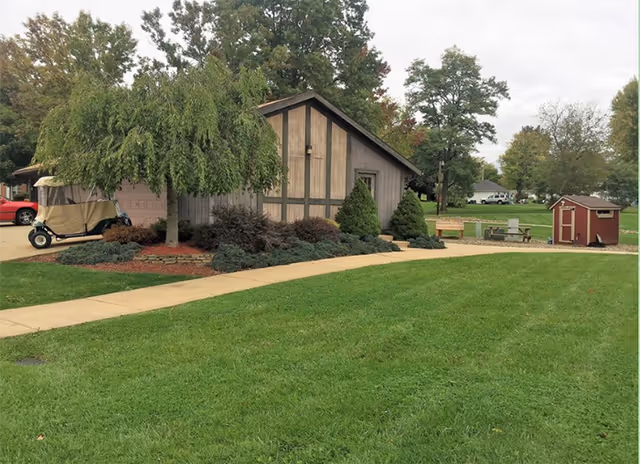 A small wood-clad building surrounded by landscaped shrubs, a paved walkway, green lawn, a parked golf cart, and a small red shed in the background.