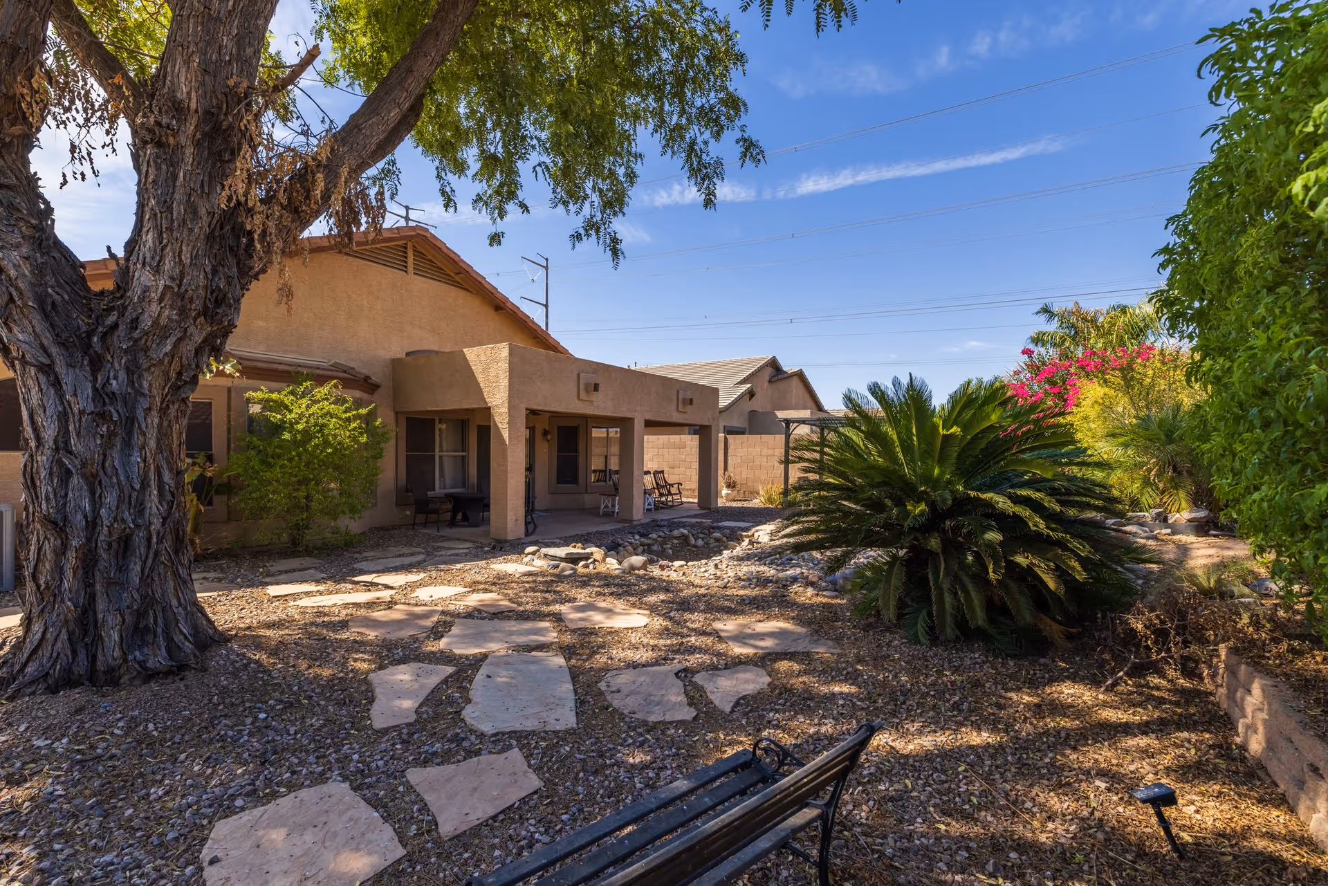 Backyard garden and covered patio of a single-story assisted living building with a stone path, trees, shrubs, and a bench.