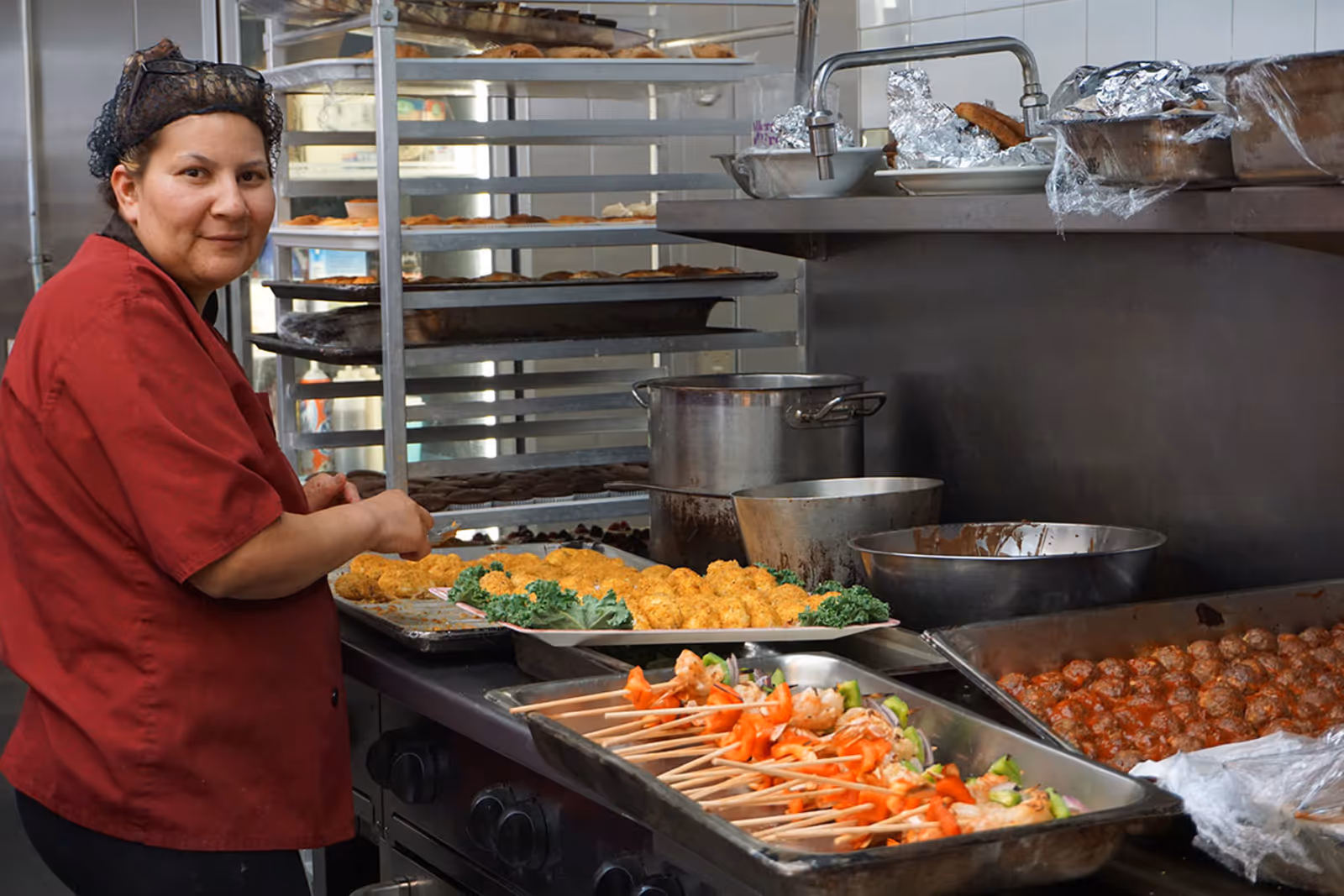 A cook in a commercial kitchen preparing trays of skewers, meatballs and breaded items on a stainless steel counter.