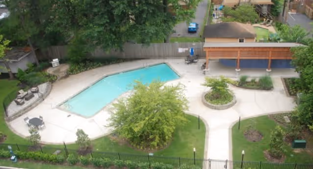 Aerial view of a fenced outdoor swimming pool with surrounding patios, seating, a pergola, and landscaped walkways.