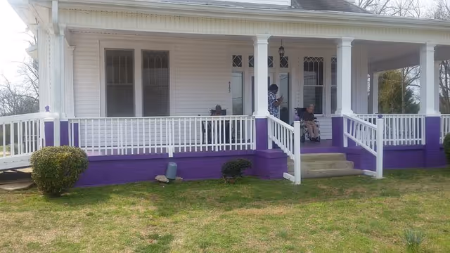 Front porch of a white house with a purple-painted foundation, white columns and railings, a ramp, and two people seated on the porch.