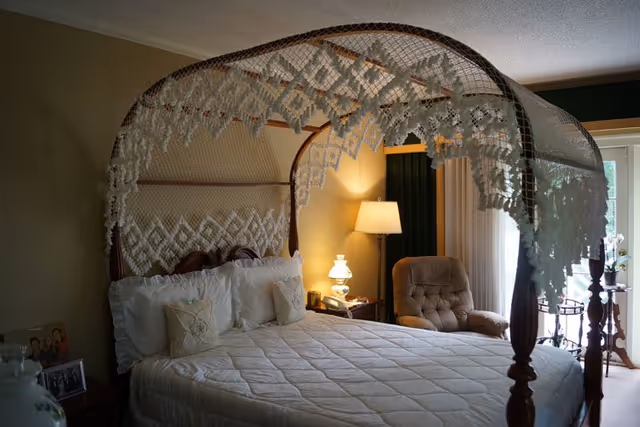 A cozy bedroom featuring a large four-poster bed with a decorative lace canopy. The bed is made with white bedding and pillows. Next to the bed is a nightstand with a lamp and a telephone. There is a comfortable armchair near a window with curtains, allowing natural light to enter the room.