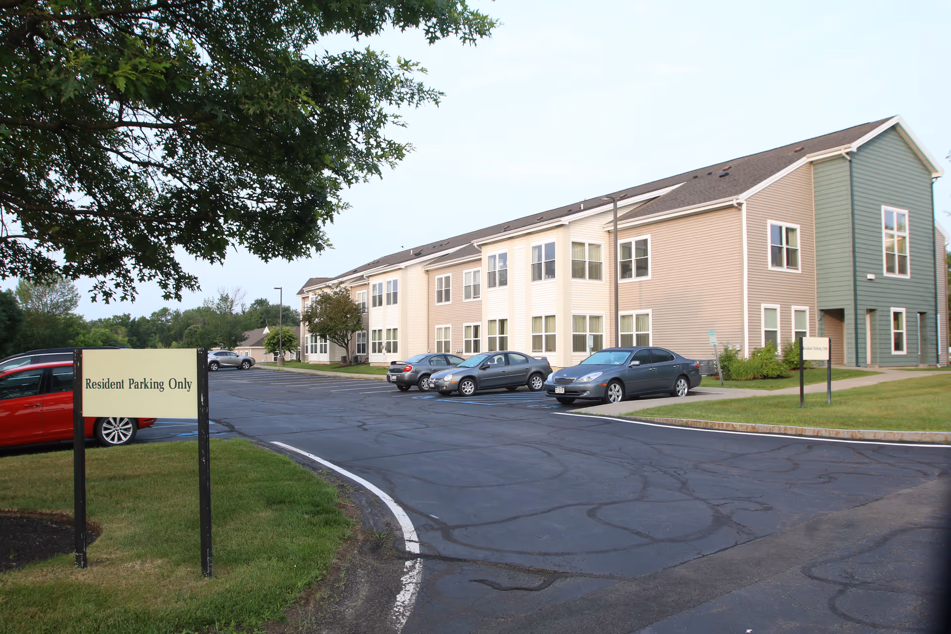 Exterior view of a two-story senior living facility building with beige and green siding. Several cars are parked in the paved parking lot in front of the building. A sign near the parking lot reads 'Resident Parking Only'. Trees and grass surround the area under a clear sky.