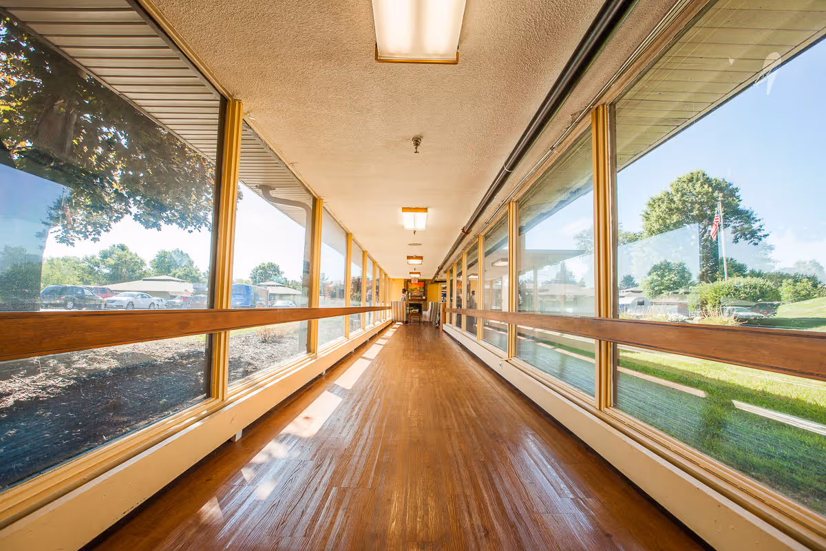 Long sunlit interior corridor with wooden floors, handrails and large windows on both sides.