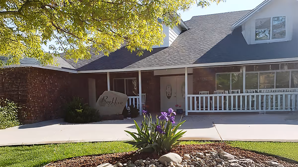 Front of a single-story senior living building with a covered porch, a stone sign reading "BeeHive", and purple irises in a flower bed.