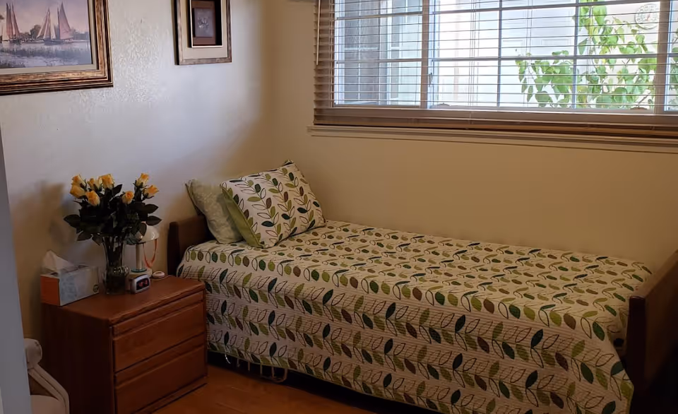 A small bedroom with a single bed dressed in green leaf-patterned bedding, a wooden nightstand with flowers, and a window with horizontal blinds.