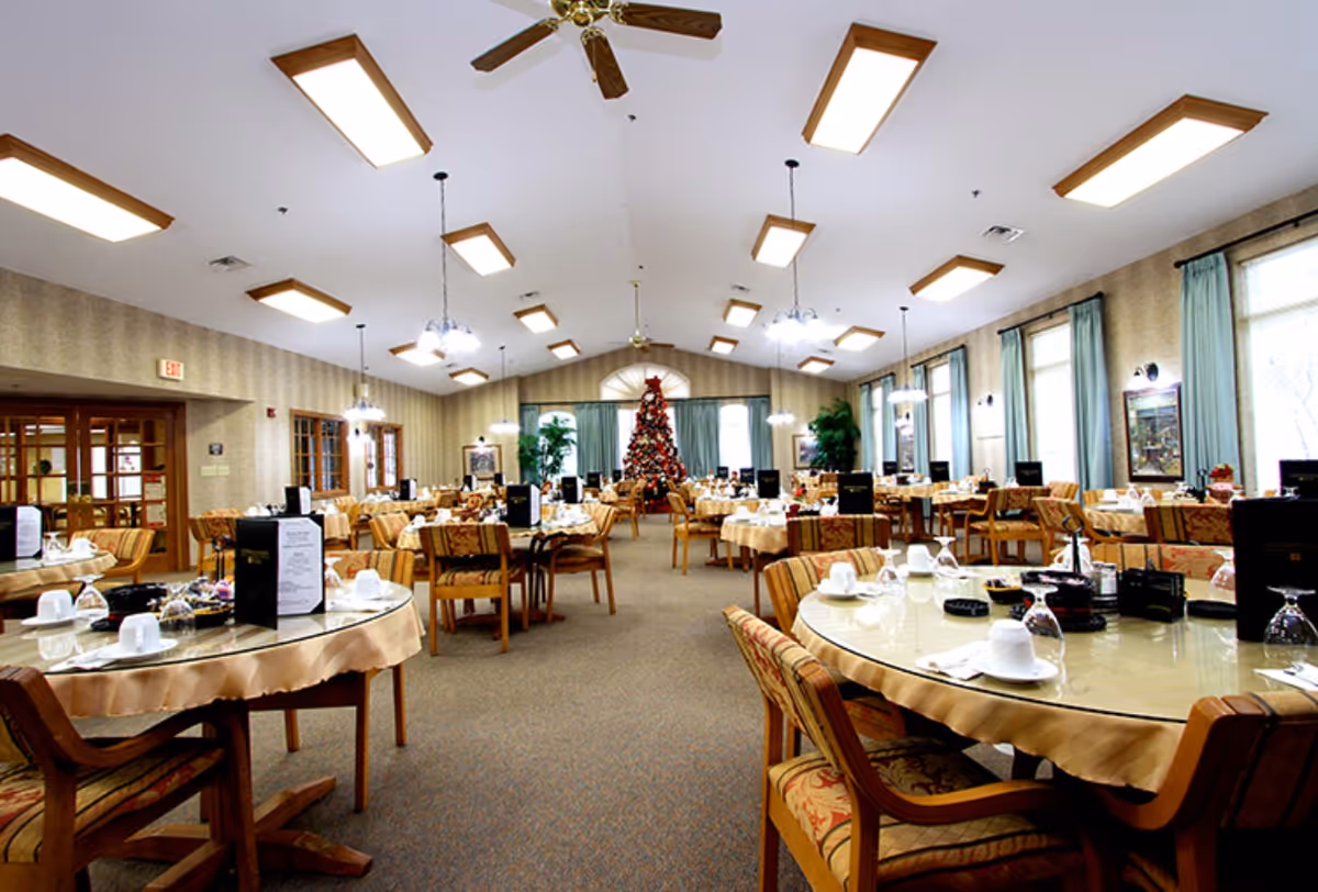 A spacious dining room with multiple round tables covered with beige tablecloths and set with white cups, glasses, and menus. The room has patterned upholstered chairs, large windows with blue curtains, ceiling fans, and bright rectangular ceiling lights. A decorated Christmas tree is visible at the far end of the room.