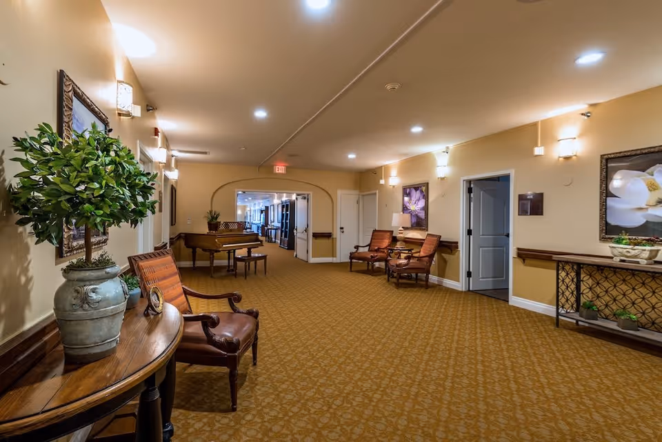 A well-lit hallway in a senior living facility with beige walls and patterned carpet. The hallway features wooden chairs with cushions, a wooden table with a potted plant, framed floral artwork on the walls, and a piano in the distance. There are wall-mounted lights and an exit sign at the far end.