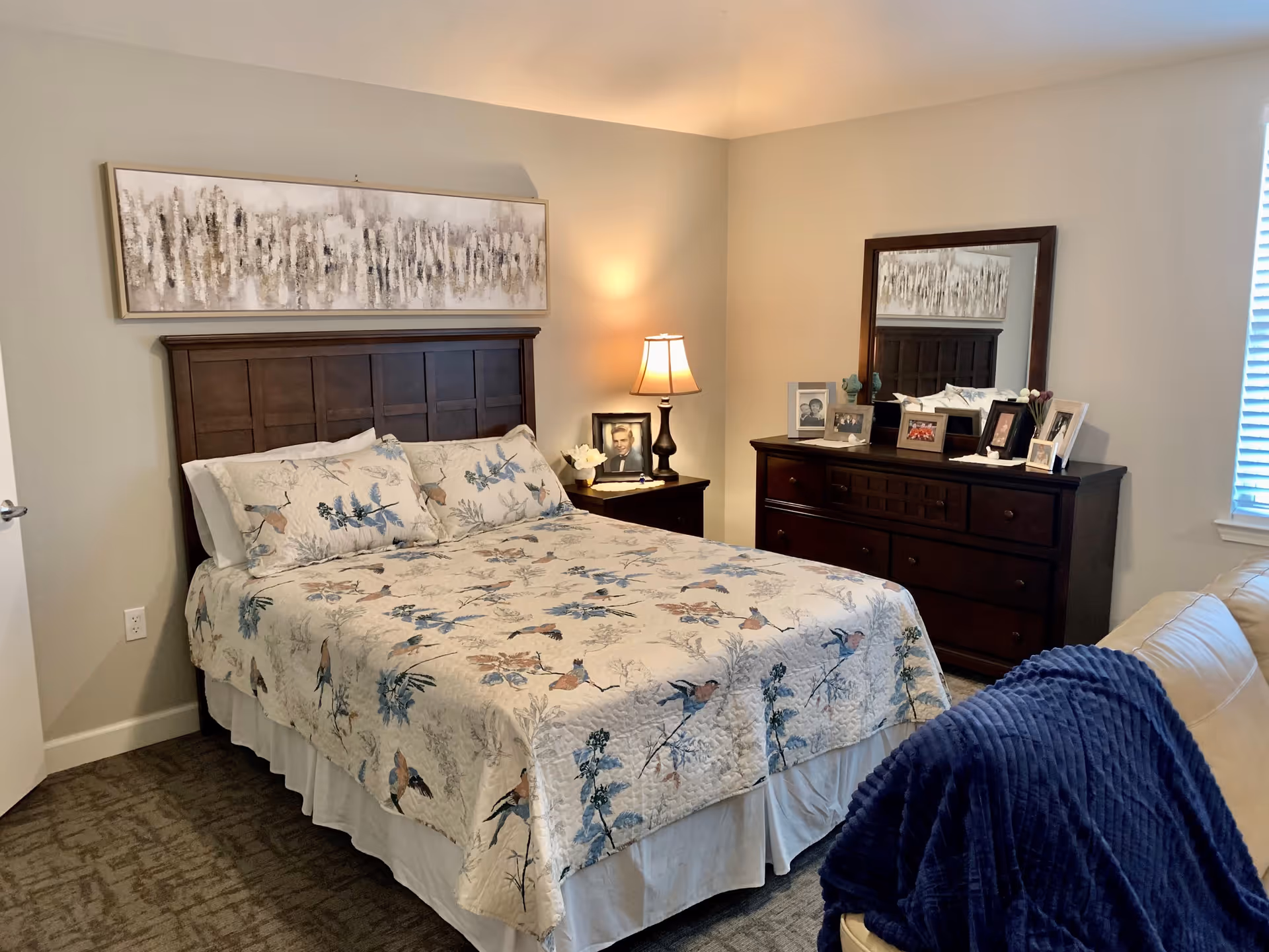 A cozy bedroom with a wooden headboard bed covered in a floral and bird patterned quilt. There is a wooden nightstand with a lamp and framed photo beside the bed. A wooden dresser with a large mirror on top holds several framed photos. A beige couch with a dark blue blanket draped over the arm is partially visible in the foreground. The room has neutral-colored walls and carpeted flooring.