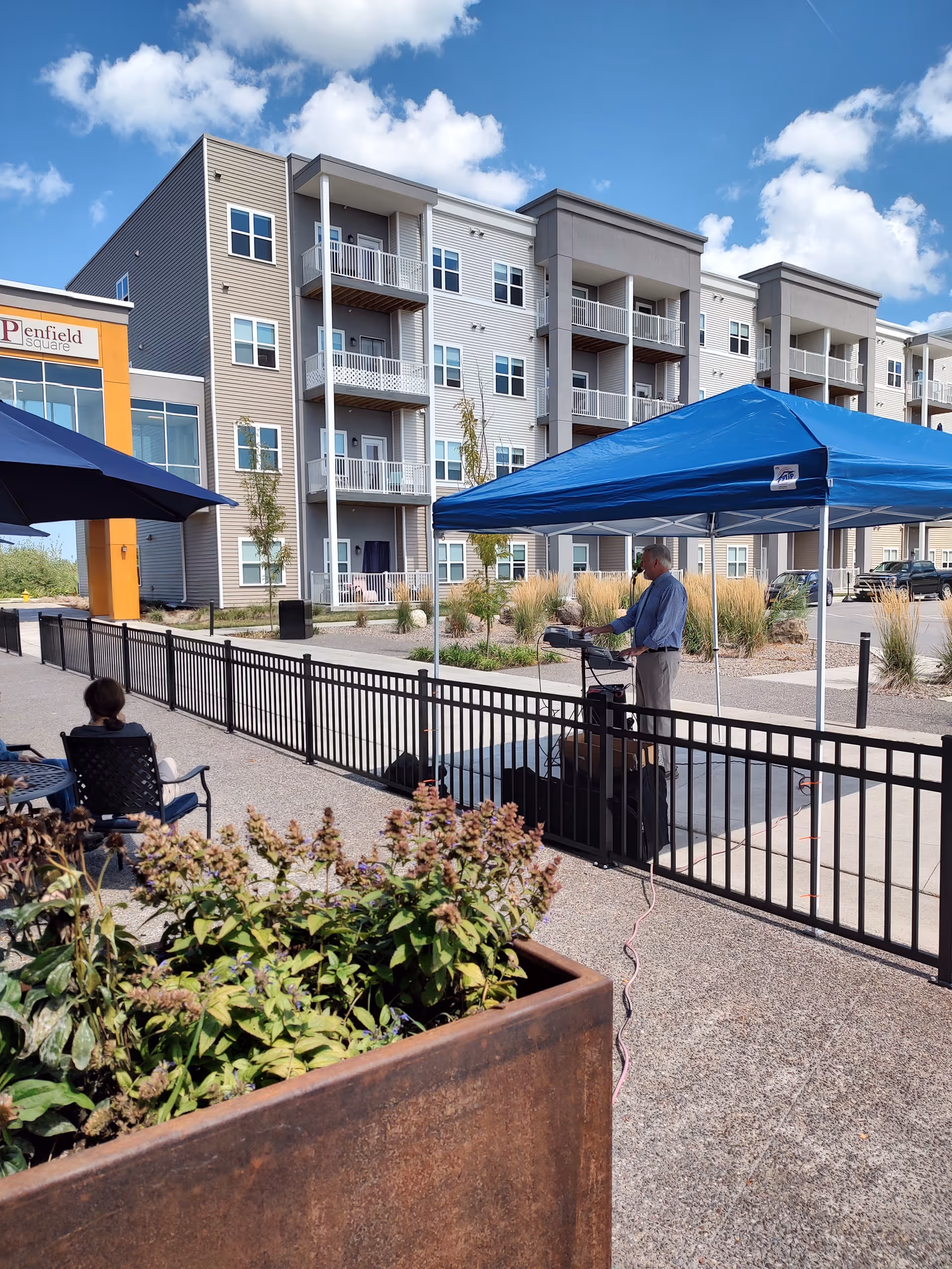Courtyard in front of a multi-story senior living building with a blue pop-up canopy and a man standing beneath it, outdoor seating, and planters in the foreground.