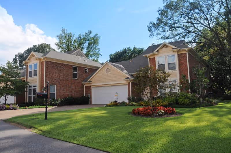 A large two-story brick house with a beige garage door, surrounded by a well-maintained green lawn and landscaped flower beds under a clear blue sky.